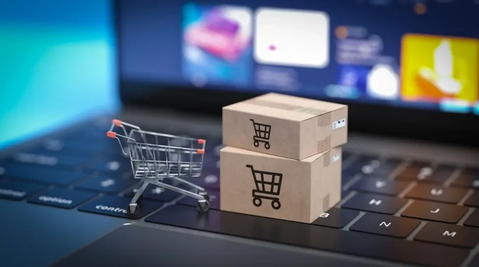 A miniature shopping cart and shipping boxes sit on the laptop of an e-commerce business owner.